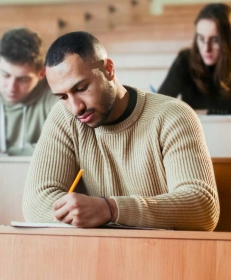 a young man in a beige sweater writing an exam in a classroom.