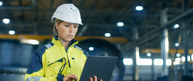 A female journeyperson in a white helmet, and yellow jacket working on a laptop while standing in a large factory with large holding tanks and equipment behind her.