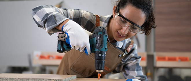 a female cabinetmaker wearing safety glasses and white gloves working with a cordless hand drill.