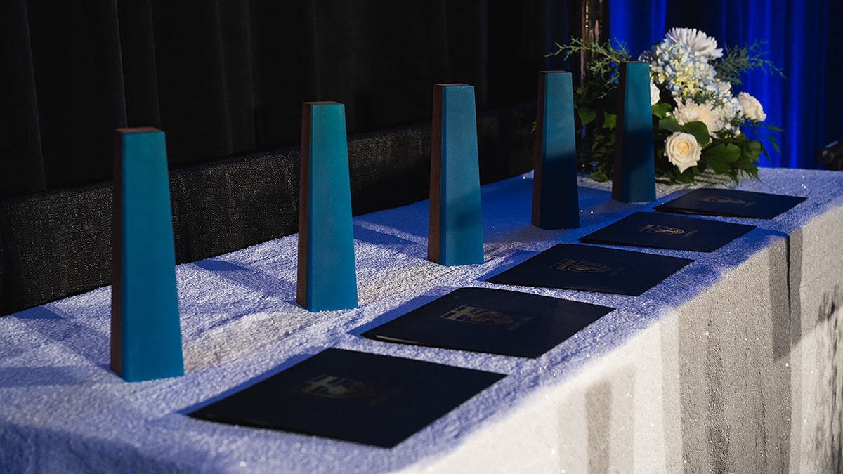 Five blue acrylic and wood Awards of Distinction trophies lined up behind certificates on a table at the award presentation evening event.