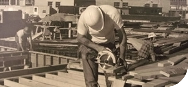 an old archived black and white photo of two male construction craft workers in safety helmets making forms for concrete footings at an outdoor construction site.