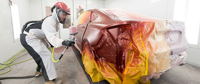 an Automotive Refinishing Technician in full white painting suit and helmet painting a vehicle in a spray booth.