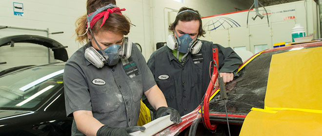 a female automotive refinishing technician journeyperson is showing a male apprentice how to block sand a vehicle.