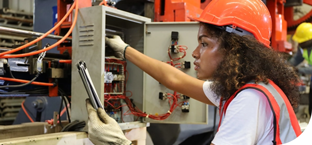 black female Industrial Electrician wearing an orange safety helmet and a safety vest working on an electrical panel in a factory.