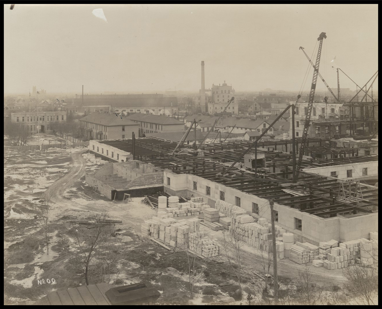 Photo with aerial view of the front of the Legislative Building under construction. There are other buildings and landmarks behind the Legislative Building and on the horizon.