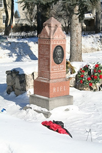 Tall obelisk headstone with an engraved portrait of Louis Riel on it. A bouquet of roses lays in the snow near the grave.