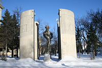 Whimsical bronze figure of Louis Riel centered in the middle of four tall, quarter-circle cement columns, which read LOUIS RIEL.
