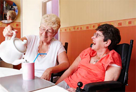 woman pouring tea from a tea pot into a cup, another woman in a wheelchair is smiling