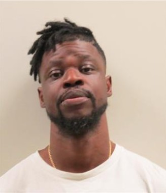 Black male standing in front of a beige wall with a white tshirt on, gold chain tucked under shirt, goatee, short hair cut on the sides and bottom and shorter dreads ontop 
